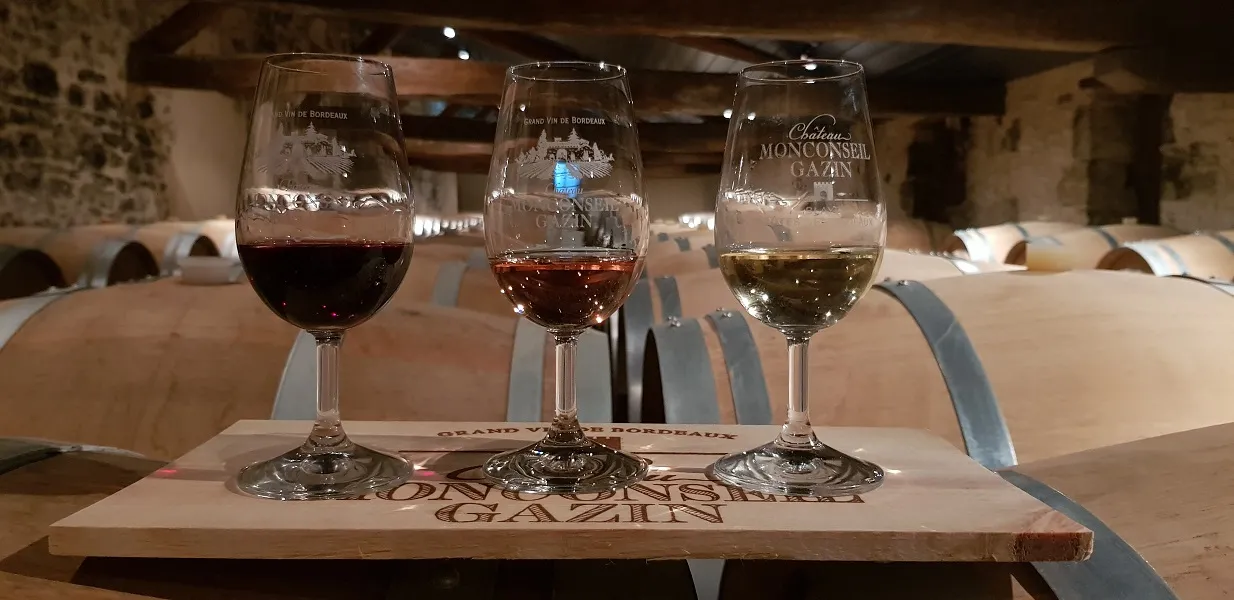 Trois verres de vin du Château Monconseil-Gazin contenant des vins de différentes couleurs, posés sur une planche en bois dans une cave avec des tonneaux en arrière-plan.