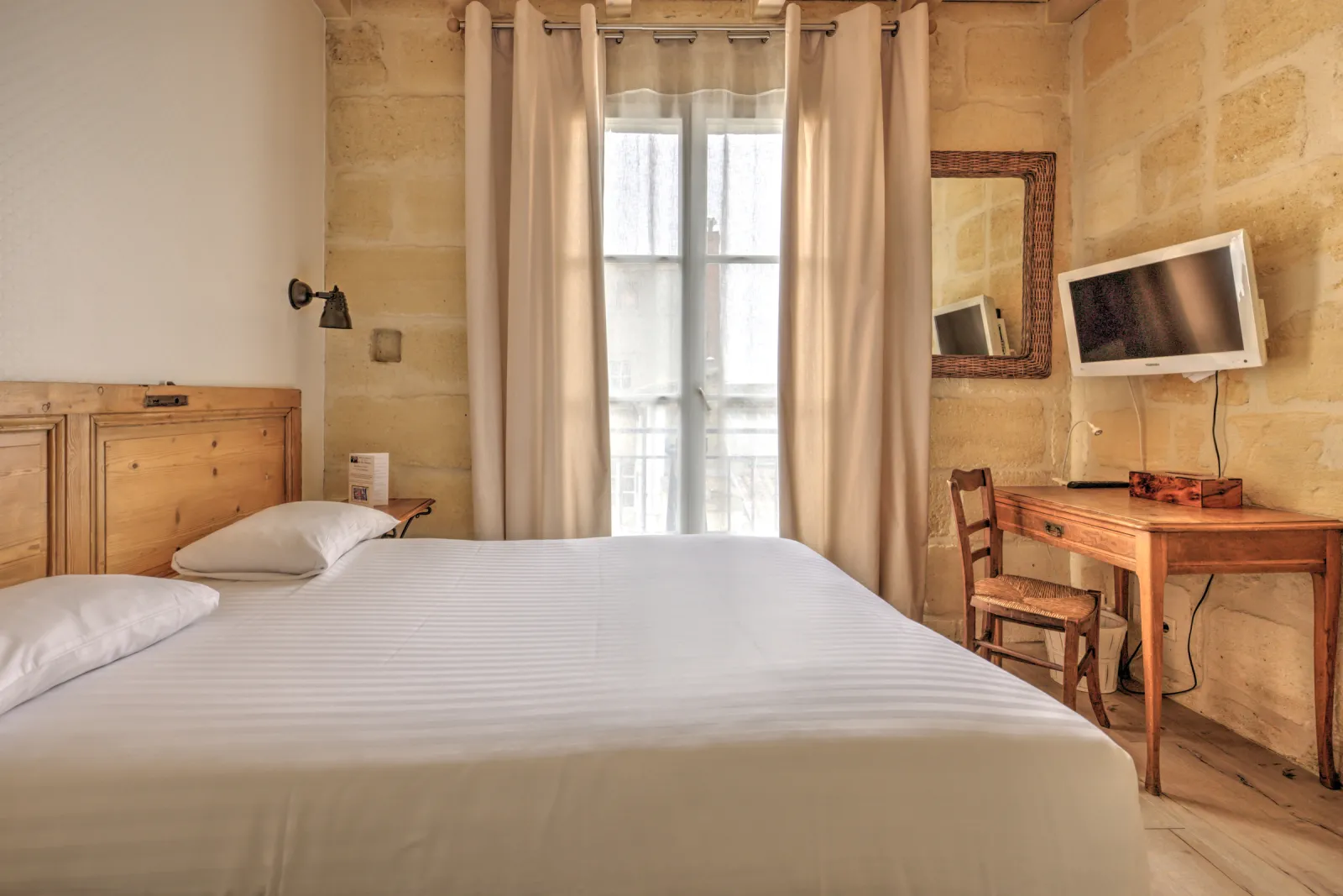 Chambre d'hôtel élégante avec lit blanc, mobilier en bois clair, bureau avec télévision, et grandes portes-fenêtres avec rideaux beige donnant sur une vue urbaine à Bordeaux.