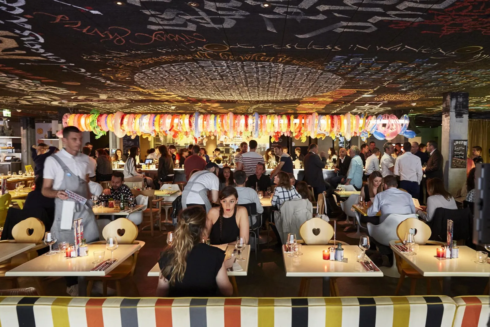 Vue intérieure du restaurant Mama Shelter à Bordeaux avec une salle bondée de clients dînant à des tables longues, décorée de lampions colorés suspendus au plafond et d'une frise à rayures multicolores en avant-plan.