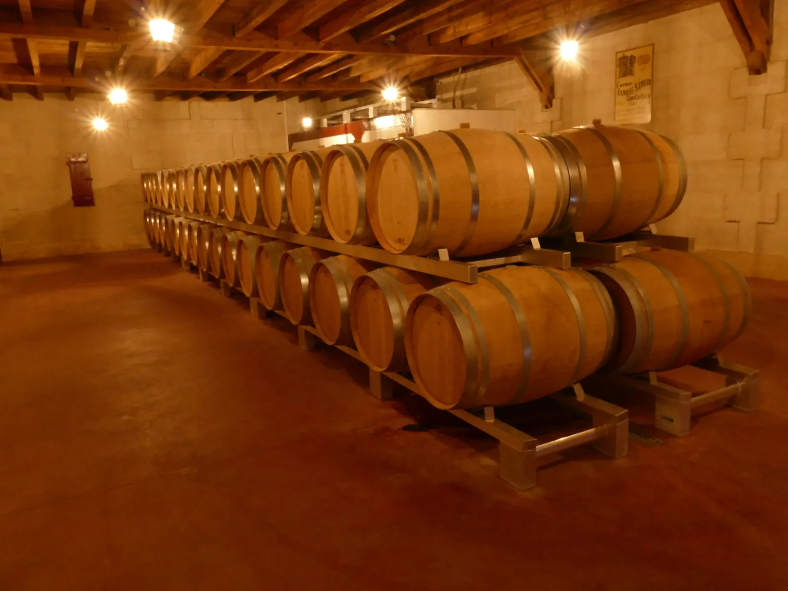 Cave de vieillement du Château Laniote à Bordeaux avec deux rangées de fûts de chêne alignés sur des supports en bois, sous un plafond à poutres apparentes avec éclairage chaleureux.