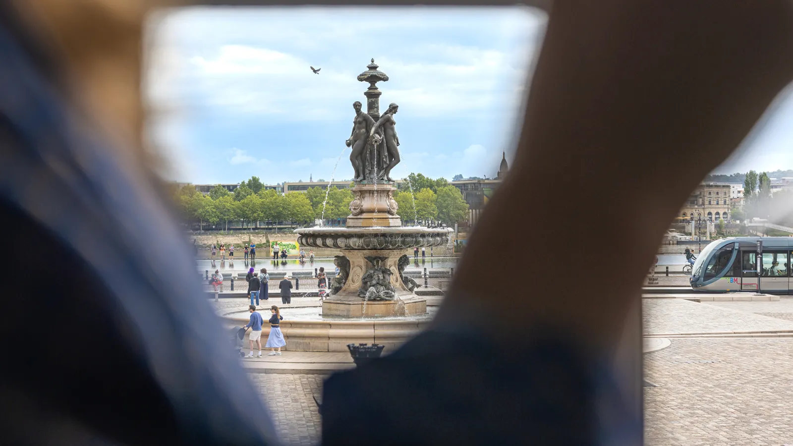 Vue encadrée d'une statue monumentale au centre d'une place publique à Bordeaux, avec des visiteurs, des bâtiments historiques et un tramway en arrière-plan