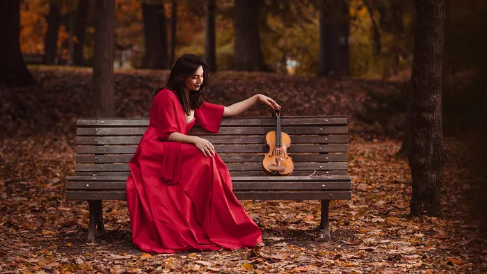 Femme en robe rouge assise sur un banc en automne, tenant un violon dans une forêt de feuilles dorées à Bordeaux