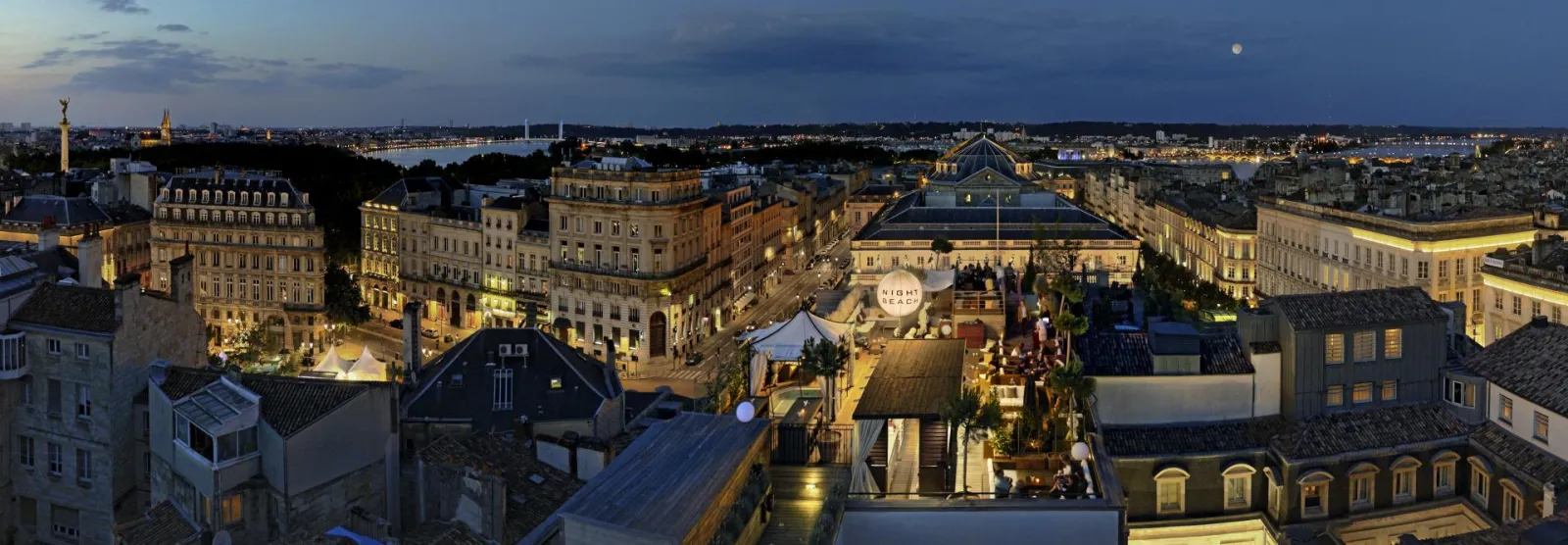 Vue aérienne crépusculaire de Bordeaux depuis la terrasse de l'Intercontinental Grand Hôtel, montrant la toit-terrasse illuminée, les bâtiments historiques dorés, et la Garonne en arrière-plan.