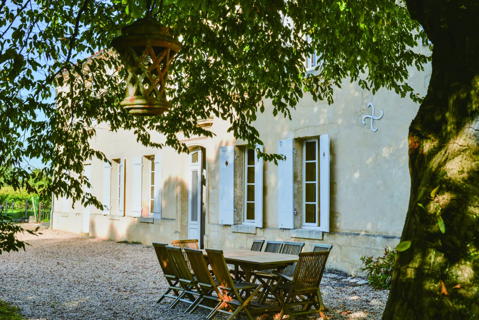 Cour ombragée du Château Valoux à Bordeaux avec une table et des chaises en bois sous un auvent, façade blanche aux volets bleus et feuillage verdoyant