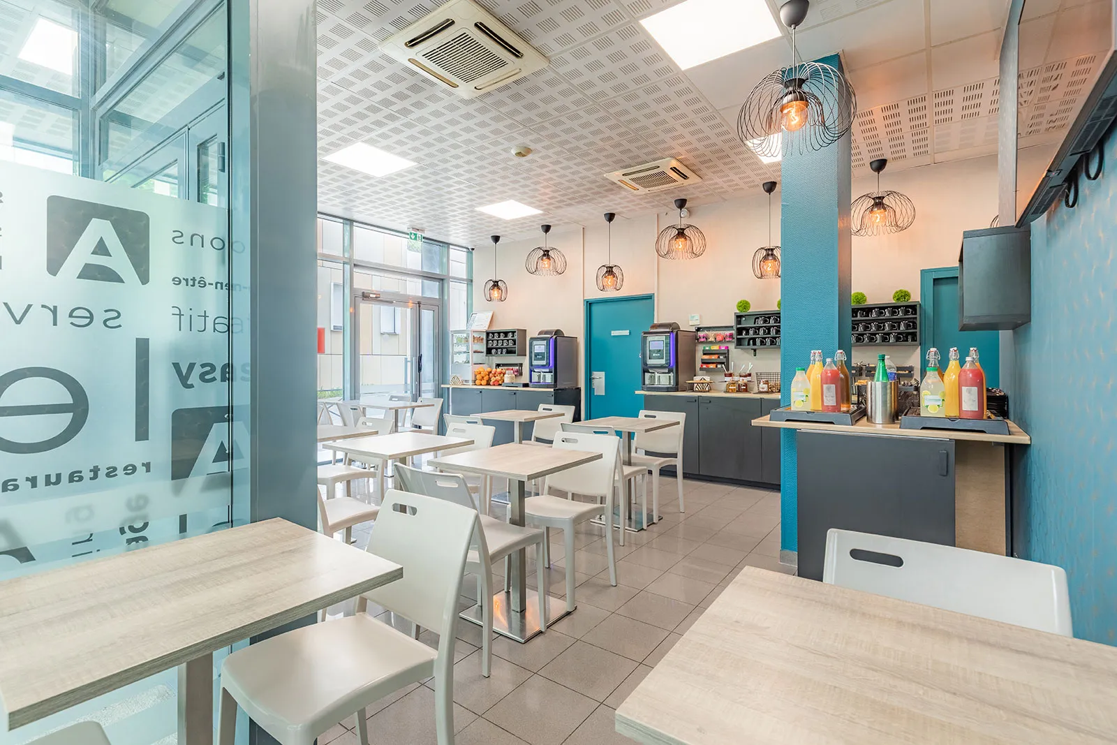 Intérieur moderne d'un café-lounge avec tables blanches, chaises beiges, comptoir turquoise, luminaires design en métal, et zone de service avec boissons et aménagements contemporains à Appart'City Classic Bordeaux Centre.