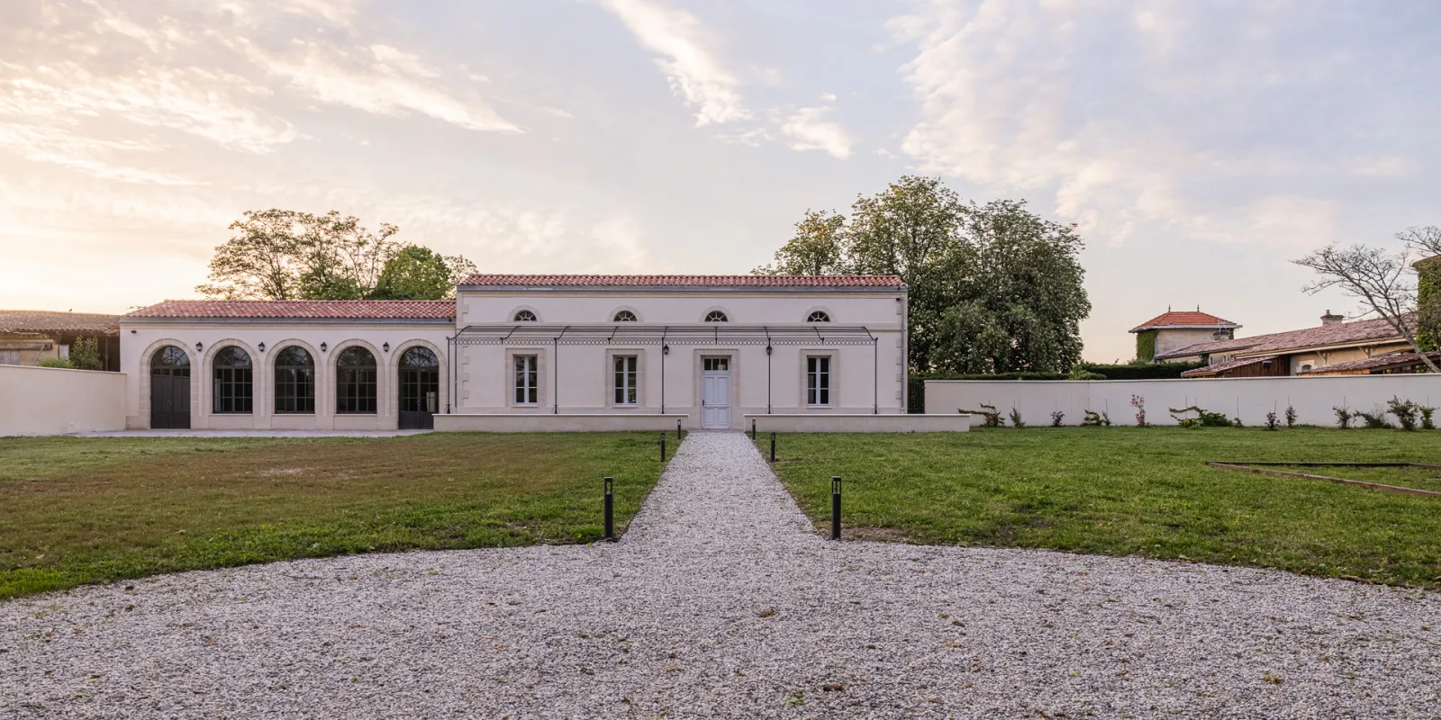 Façade blanche du Château Sénéjac à Bordeaux avec allée de gravier, pelouses vertes et toiture en tuiles rouges, entouré de murs blancs et d'arbres.
