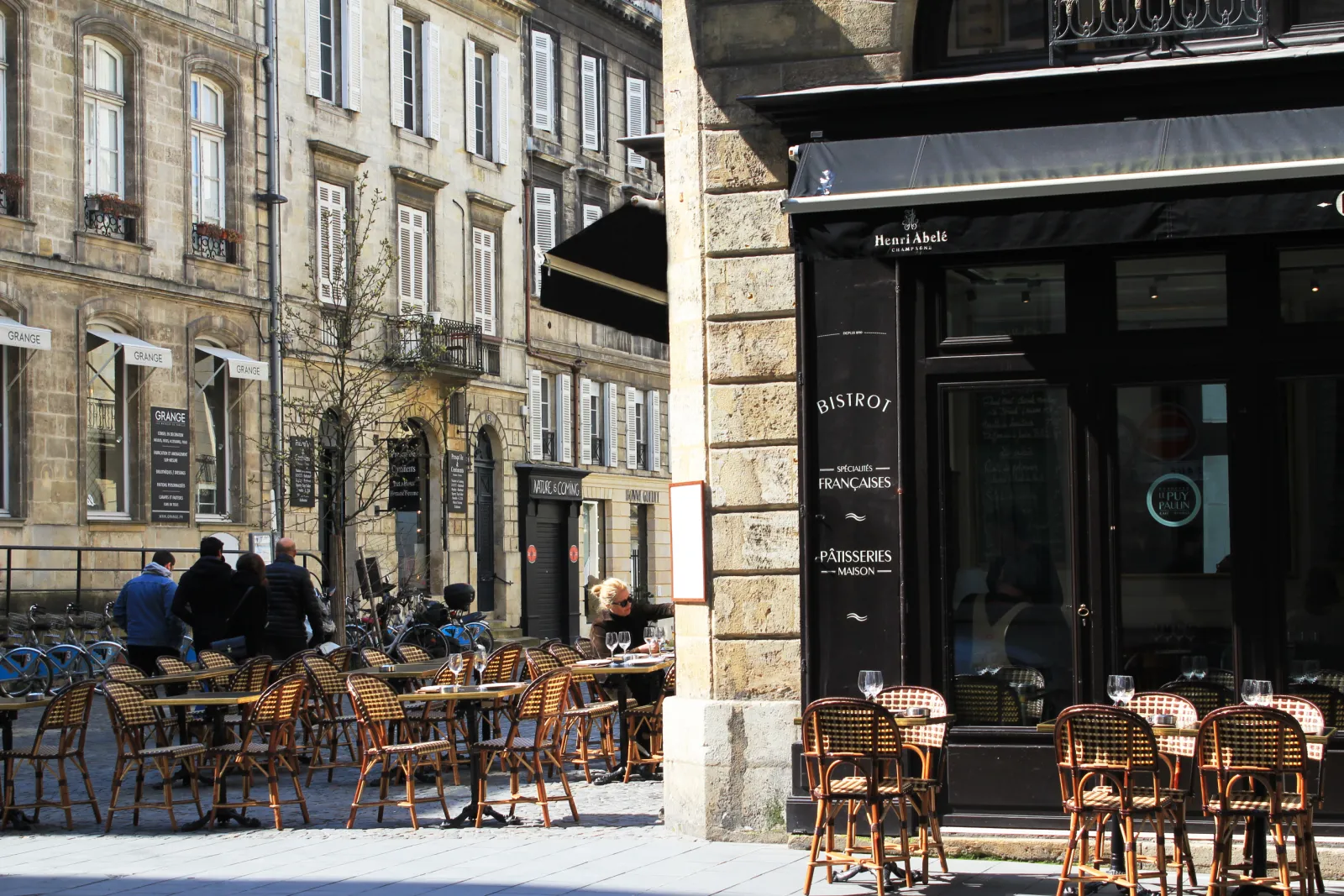 Terrasse de restaurant avec chaises en rotin face à un bistrot parisien aux façades en pierre, avec des clients assis et des bâtiments haussmanniens en arrière-plan à Bordeaux