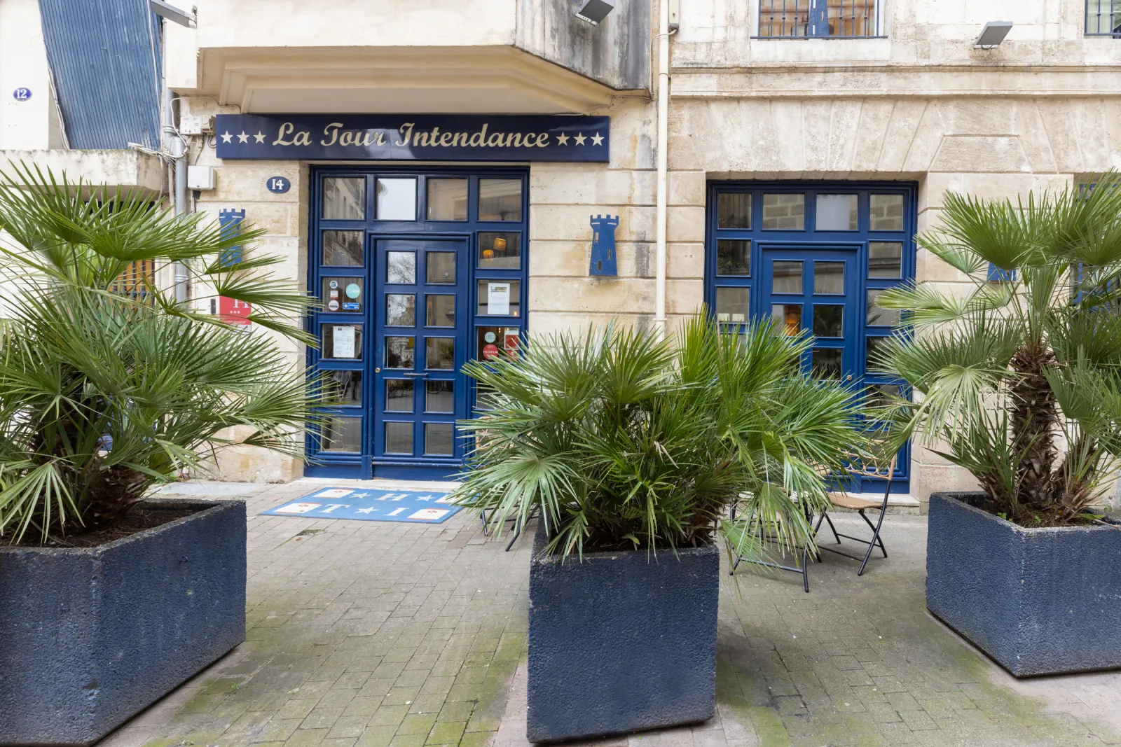 Façade de l'Hôtel de la Tour Intendance à Bordeaux avec portes bleues, enseigne navy et blanc, planteurs de palmiers en façade, architecture de pierre classique
