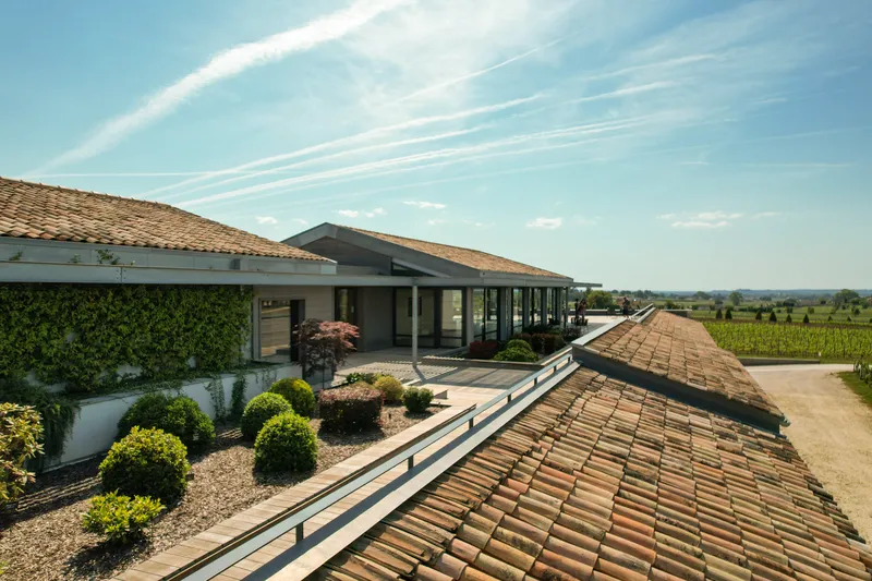 Vue panoramique du Château Balestard La Tonnelle à Bordeaux, montrant une maison moderne aux toits de tuiles brunes avec terrasse, entourée de vignobles verdoyants sous un ciel bleu dégagé.