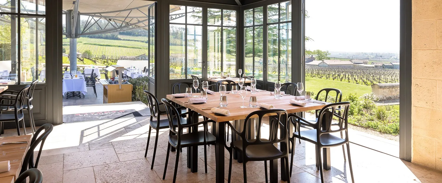 Salle de restaurant élégante avec grandes baies vitrées donnant sur les vignobles de Bordeaux, table en bois avec chaises noires, verres et couverts, vue panoramique sur les vignes et paysage rural.