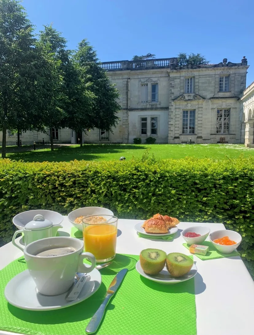 Petit-déjeuner élégant servi en terrasse avec vue sur le Château La Chèze à Bordeaux, comprenant café, jus d'orange, croissant, kiwis et accompagnements sur nappes vertes.