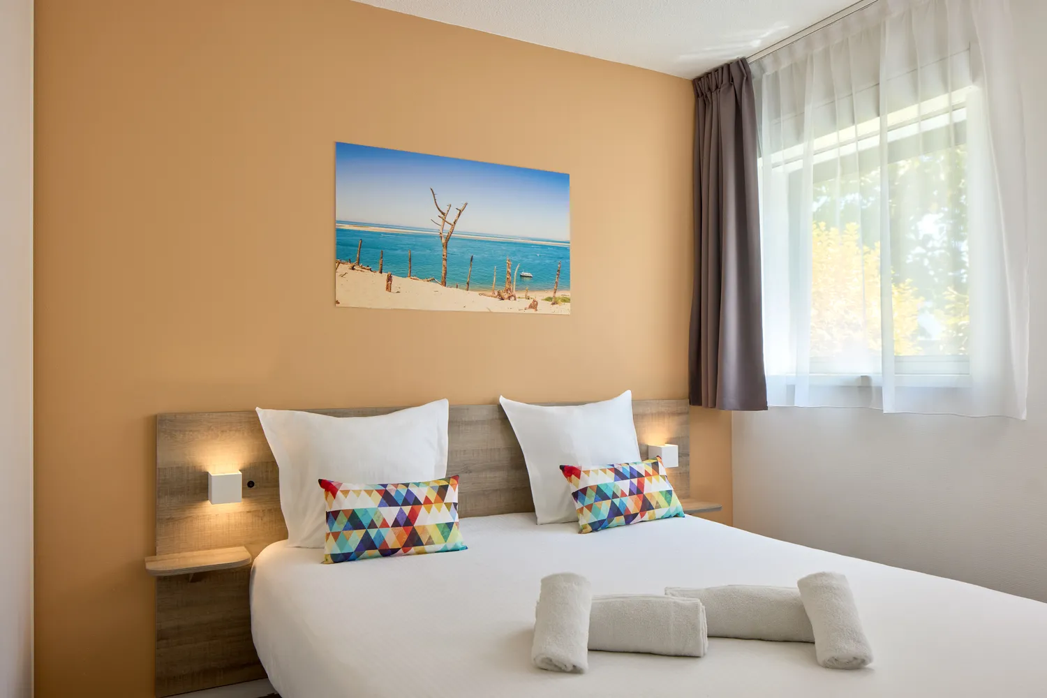 Chambre moderne avec lit double, murs beige clair, tableau de plage au-dessus du lit, oreillers colorés géométriques, deux serviettes blanches roulées, lampes de chevet intégrées, fenêtre avec double rideau et vue sur la verdure.