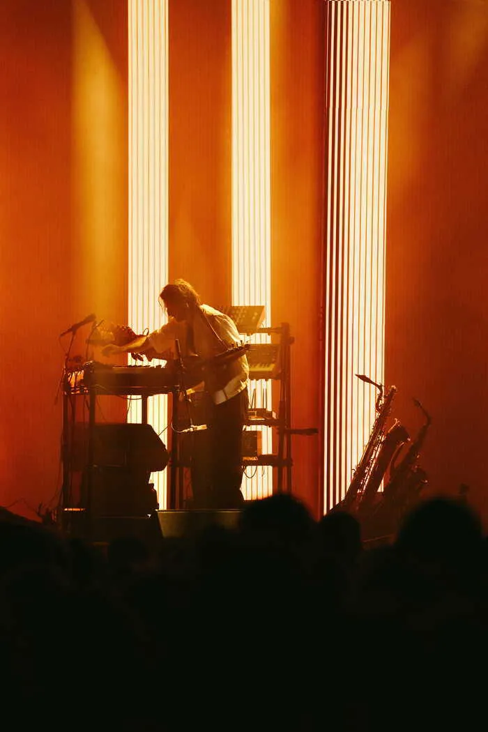 Concert de Thylacine à Bordeaux: musicien jouant du clavier sur scène avec éclairage orange et colonnes lumineuses, saxophoniste en arrière-plan, silhouettes du public au premier plan.