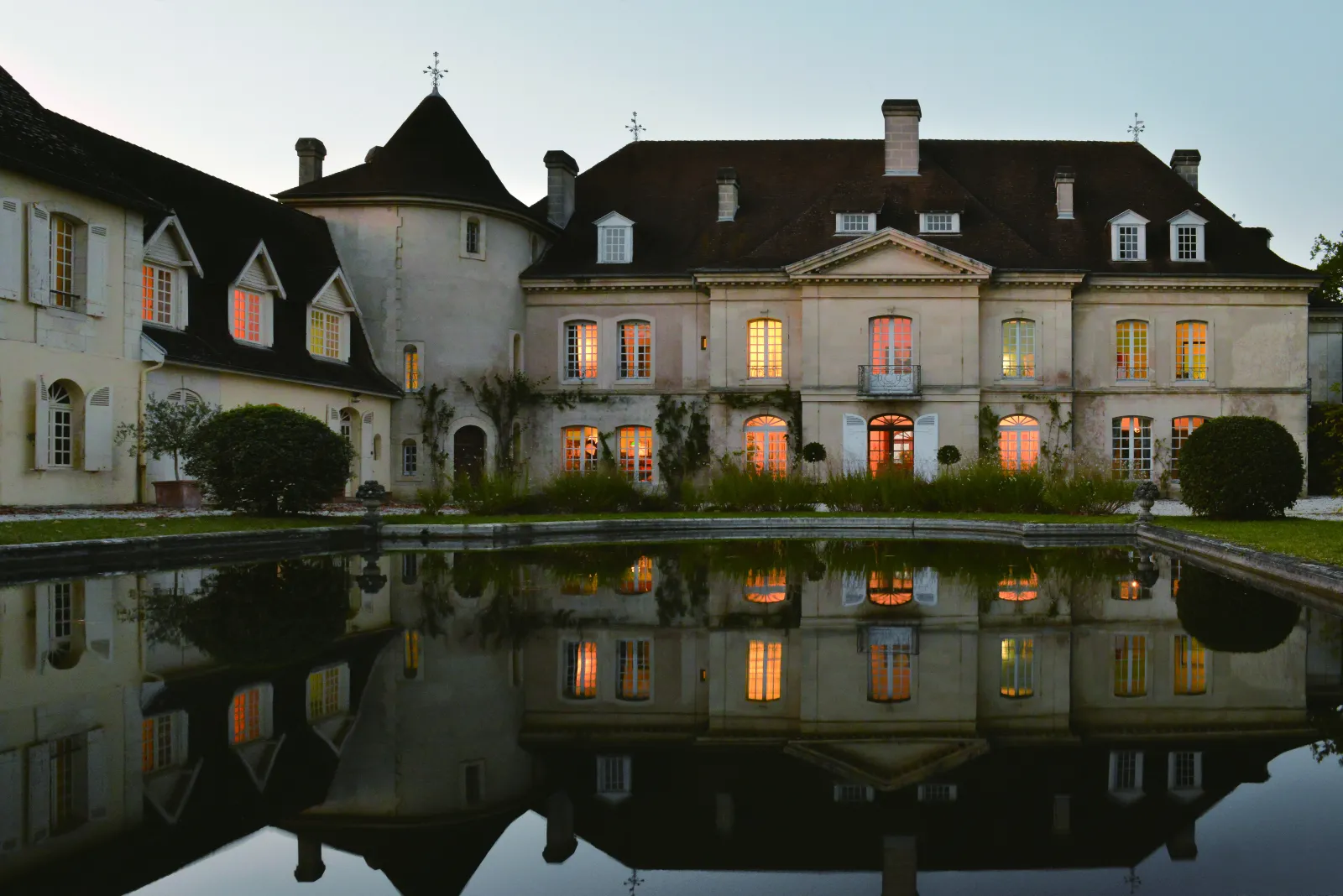 Château Bouscaut à Bordeaux illuminé au crépuscule, façade classique avec fenêtres éclairées se reflétant dans le bassin d'eau devant le bâtiment.