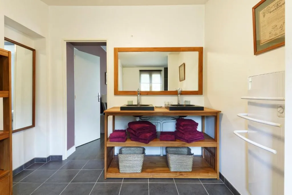 Salle de bain moderne avec double vasque sur meuble en bois, miroir rectangulaire encadré, serviettes burgundy empilées, paniers de rangement, carrelage gris et portes en bois à la Bastide de Siran, Bordeaux