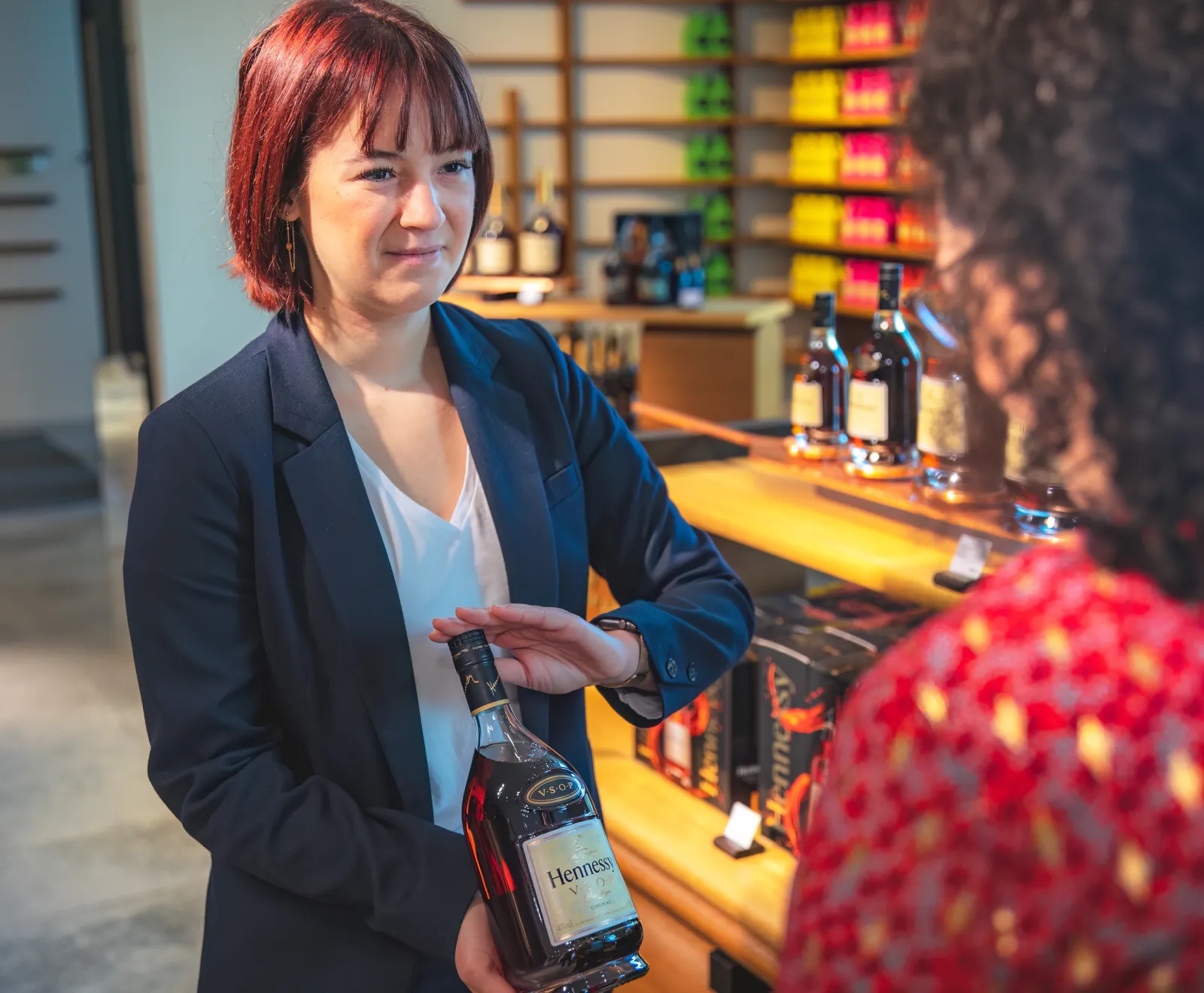 Une femme aux cheveux roux présente une bouteille de Hennessy dans la distillerie de Bordeaux, face à un client lors d'une visite guidée.