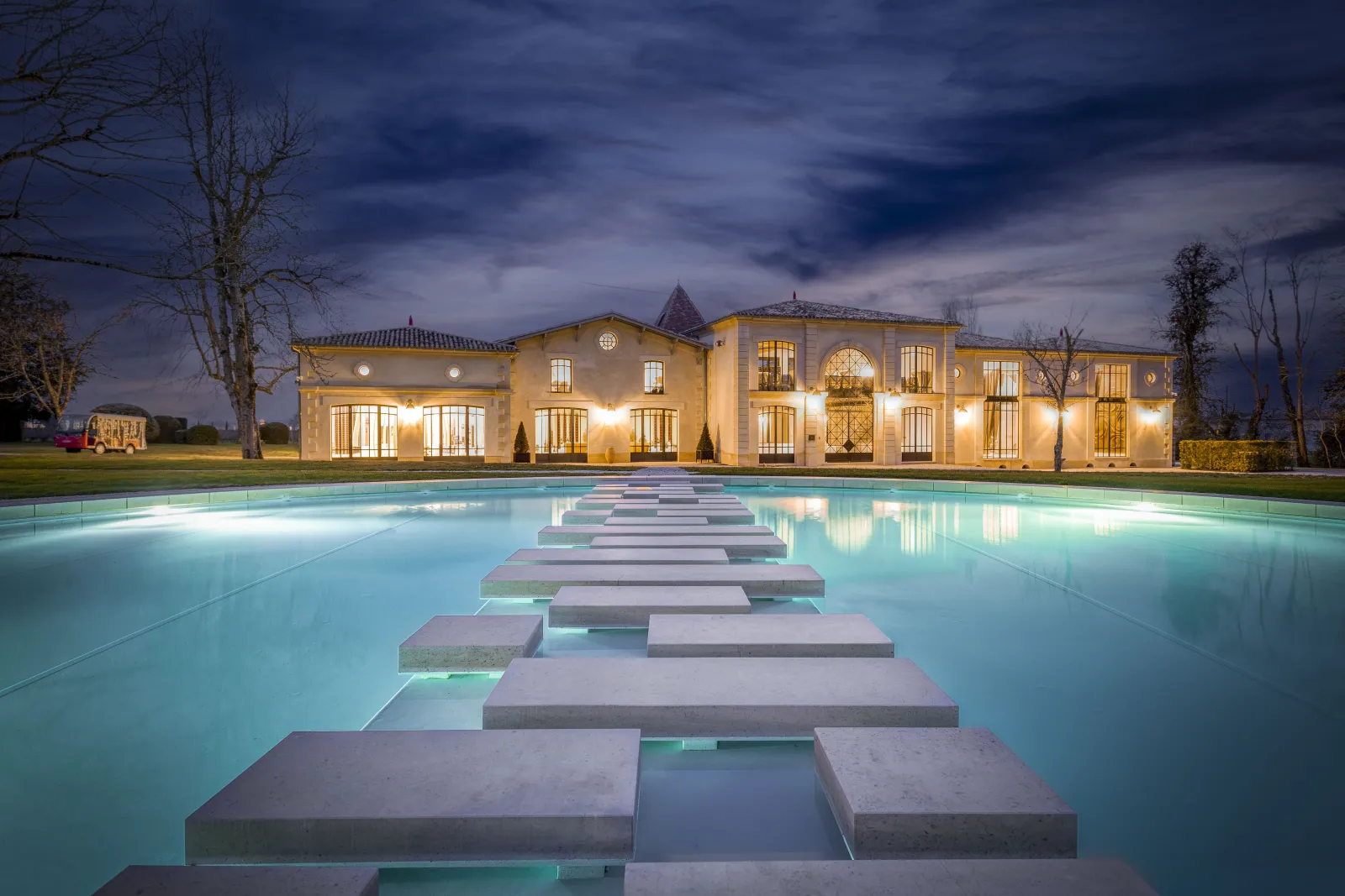 Vue nocturne du Château Montlabert à Bordeaux avec sa façade illuminée en jaune, surplombant une piscine moderne aux eaux turquoise et dalles de pierre alignées en perspective.