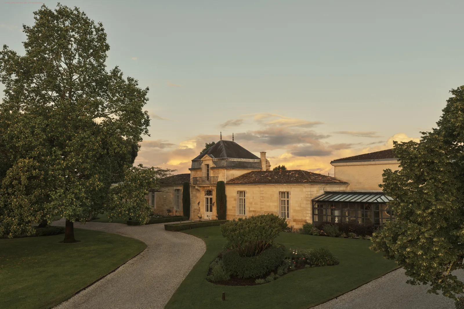 Villa Cordeillan-Bages à Bordeaux au coucher du soleil, avec façade en pierre claire, toit mansardé, allée gravillonnée et jardins paysagers encadrés de grands arbres.
