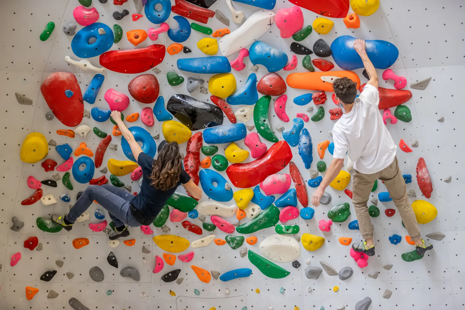 Deux personnes escaladent un mur d'escalade intérieur coloré parsemé de prises multicolores (bleues, rouges, vertes, jaunes, orange et roses) à la station UCPA Sport de Bordeaux.