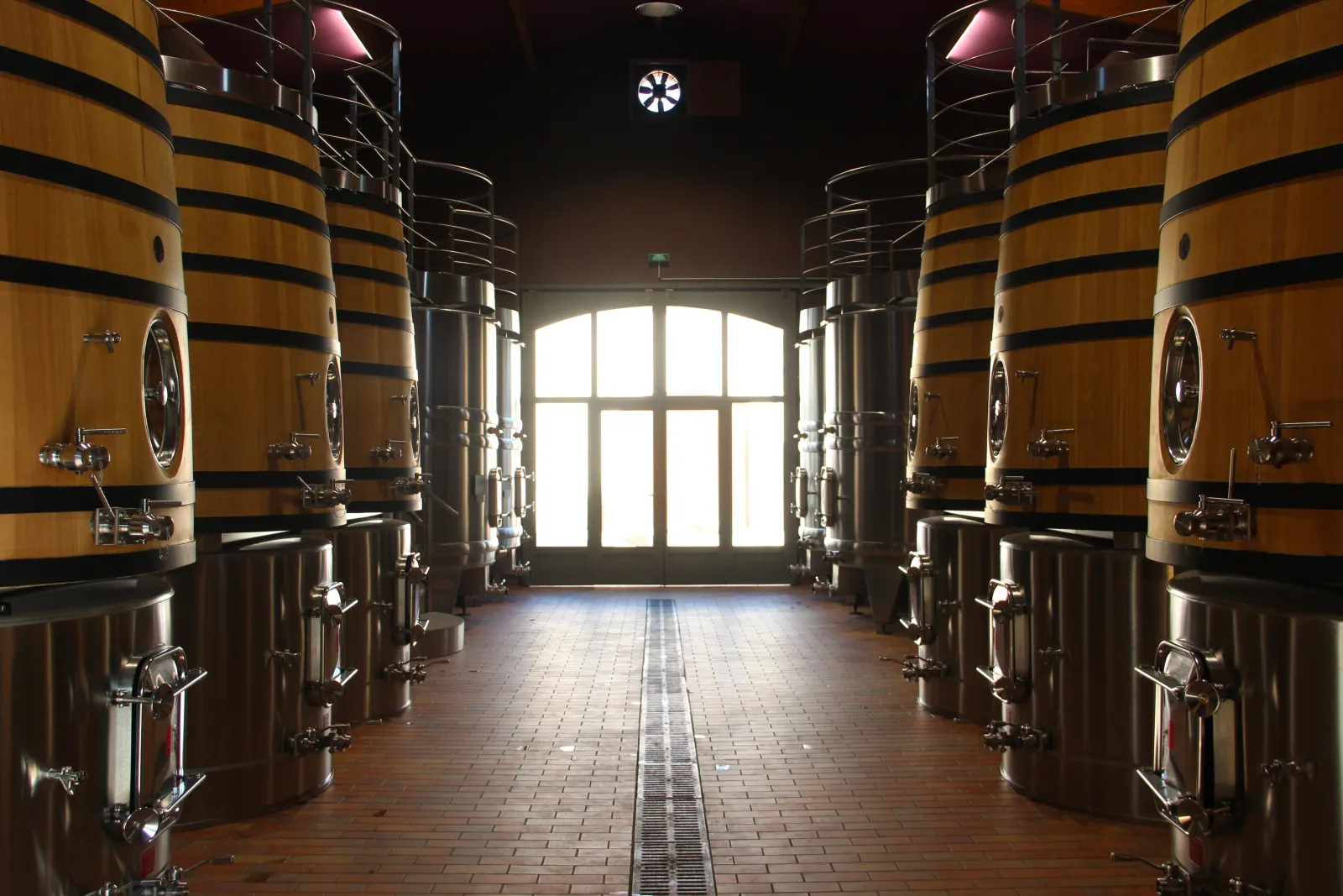 Intérieur d'une cave vinicole avec des tonneaux en bois cerclés de métal alignés de chaque côté d'une allée centrale, menant à une porte vitrée au fond, au Château Bernateau à Bordeaux.