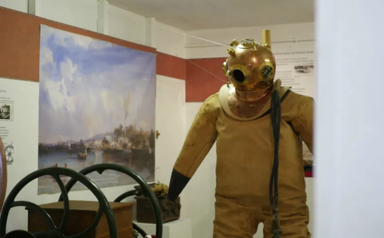 Mannequin portant un scaphandre de plongée ancien en laiton doré avec casque sphérique, exposé au Musée des Amis du Vieux Lormont à Bordeaux, à côté d'une roue de navire et d'une peinture maritime.