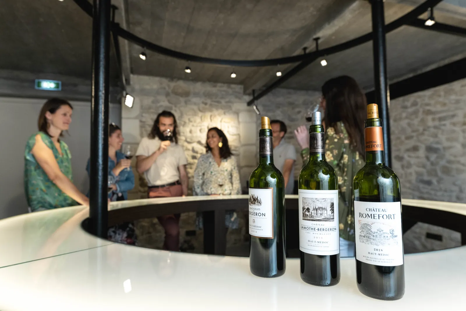 Trois bouteilles de vin du Château Romefort et Château Lamothe Bergeron exposées sur un comptoir blanc, avec des visiteurs en arrière-plan découvrant les vins dans une cave de Bordeaux.