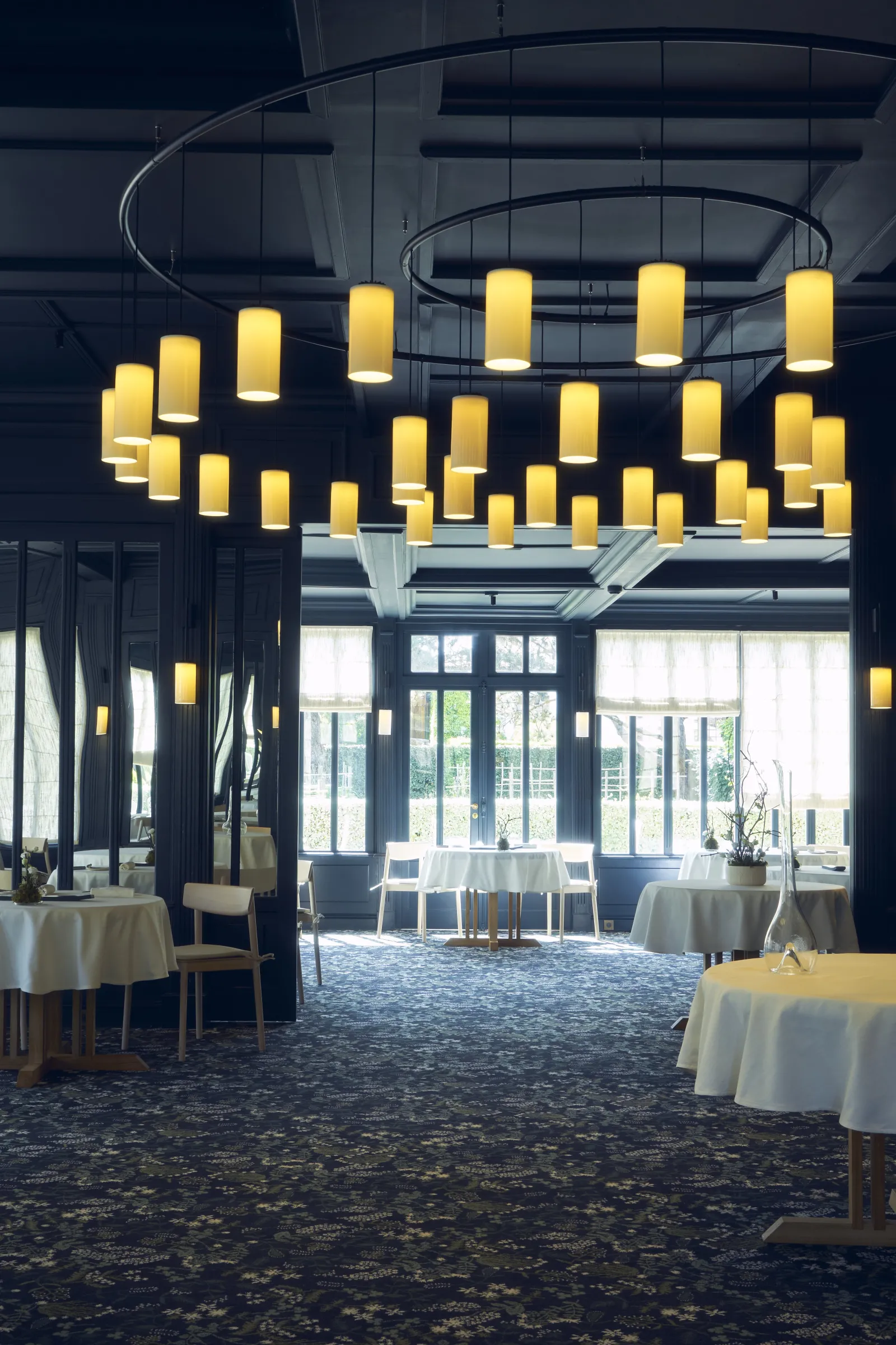 Salle de restaurant élégante avec lustre moderne composé de cylindres lumineux jaunes, tables rondes recouvertes de nappes blanches, grandes baies vitrées, plafond architectural en courbes et colonnes grises, à La Grand'Vigne à Bordeaux.