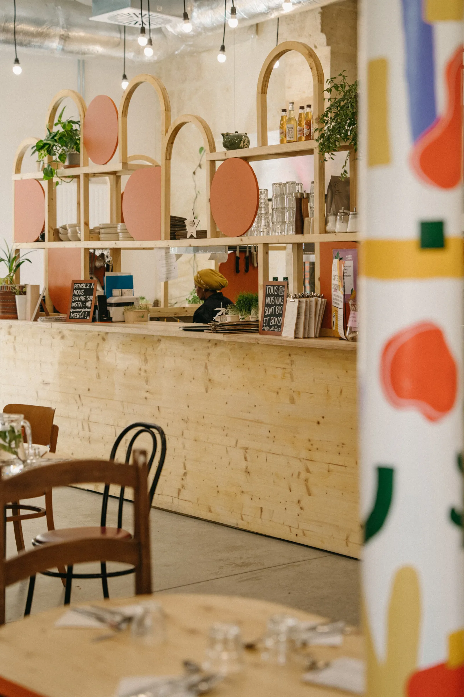 Intérieur moderne du restaurant Marie Curry à Bordeaux avec comptoir en bois clair, étagères géométriques aux formes arrondies peint en tons pastel, miroirs circulaires rose poudré, plantes vertes, chaises vintage marron, et détails colorés muraux.
