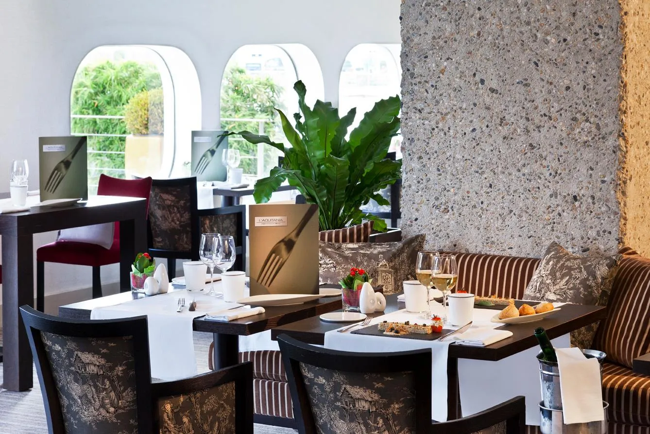 Restaurant élégant L'Aquitania à Bordeaux avec tables dressées, chaises ornées de motifs floraux, grande baie vitrée arquée, plantes vertes et murs en béton brut.