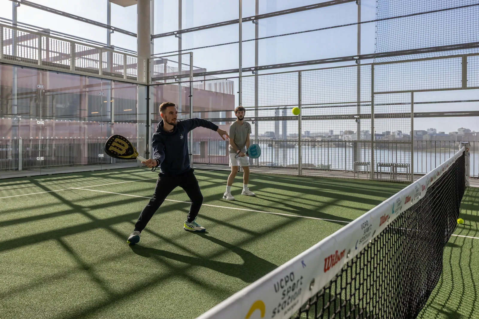 Deux joueurs de padel en action sur un court intérieur vitré à la UCPA Sport Station de Bordeaux, avec vue sur la Garonne et la ville en arrière-plan.