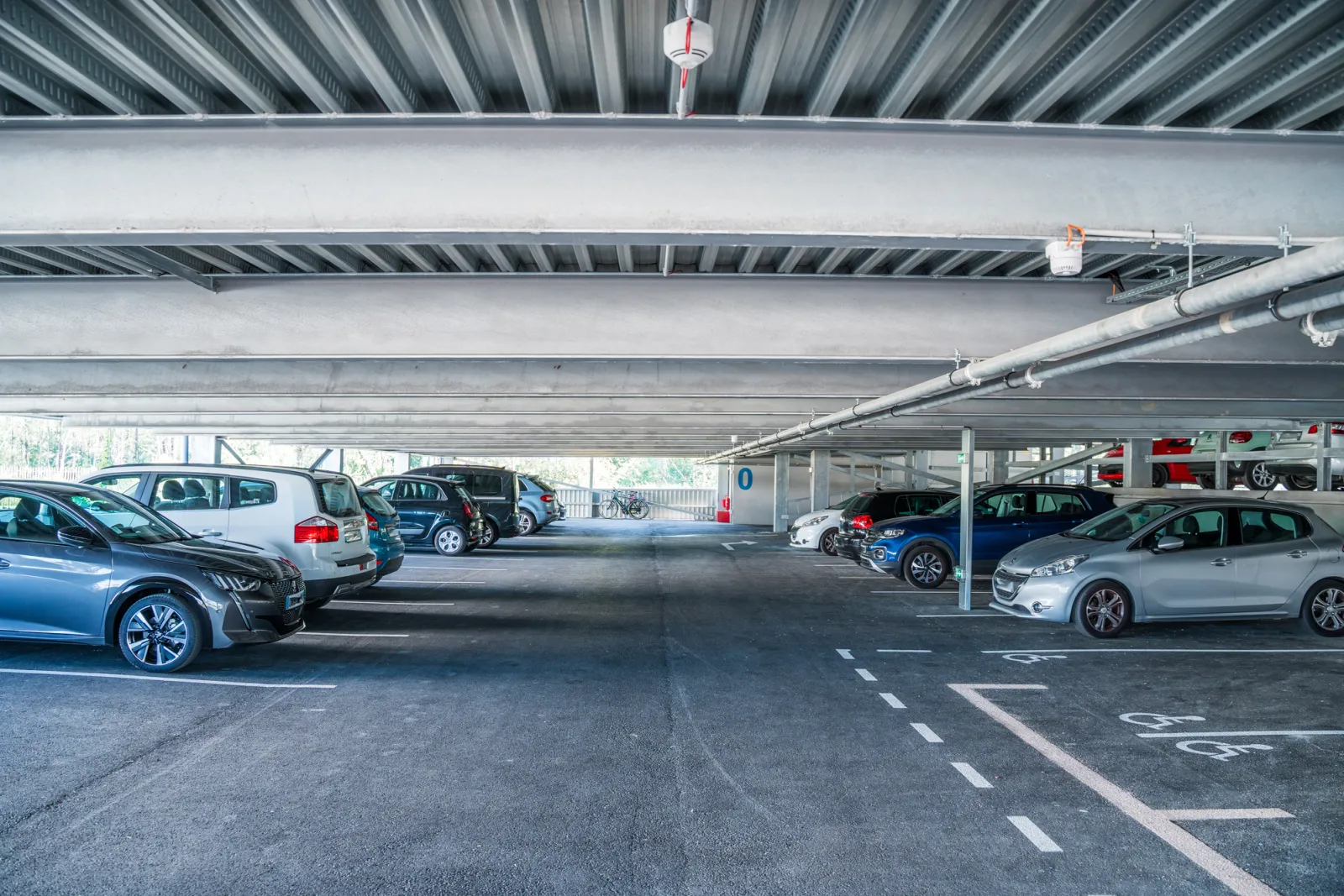 Parking couvert souterrain avec plusieurs véhicules garés sous une structure en béton avec poutres et installations de ventilation.