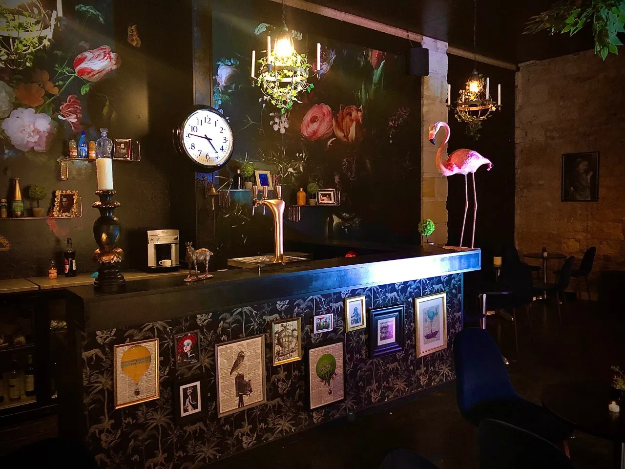 Intérieur élégant du Théâtre des Chartrons à Bordeaux avec un comptoir de bar noir aux façades décorées, murs verts ornés de fleurs roses, horloge blanche, lustre doré, flamant rose néon et éléments de décoration vintage.