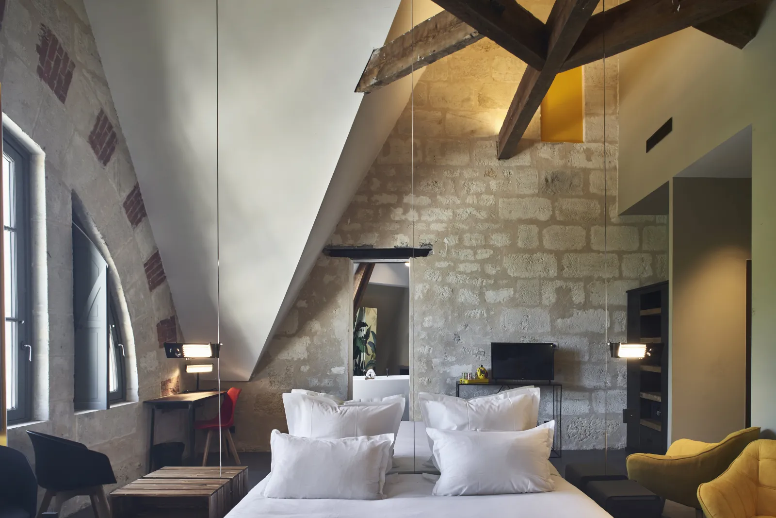 Chambre mansardée moderne de l'Hôtel La Zoologie à Bordeaux, avec lit blanc, miroir, murs en pierre brute, poutres apparentes et accents de couleur jaune