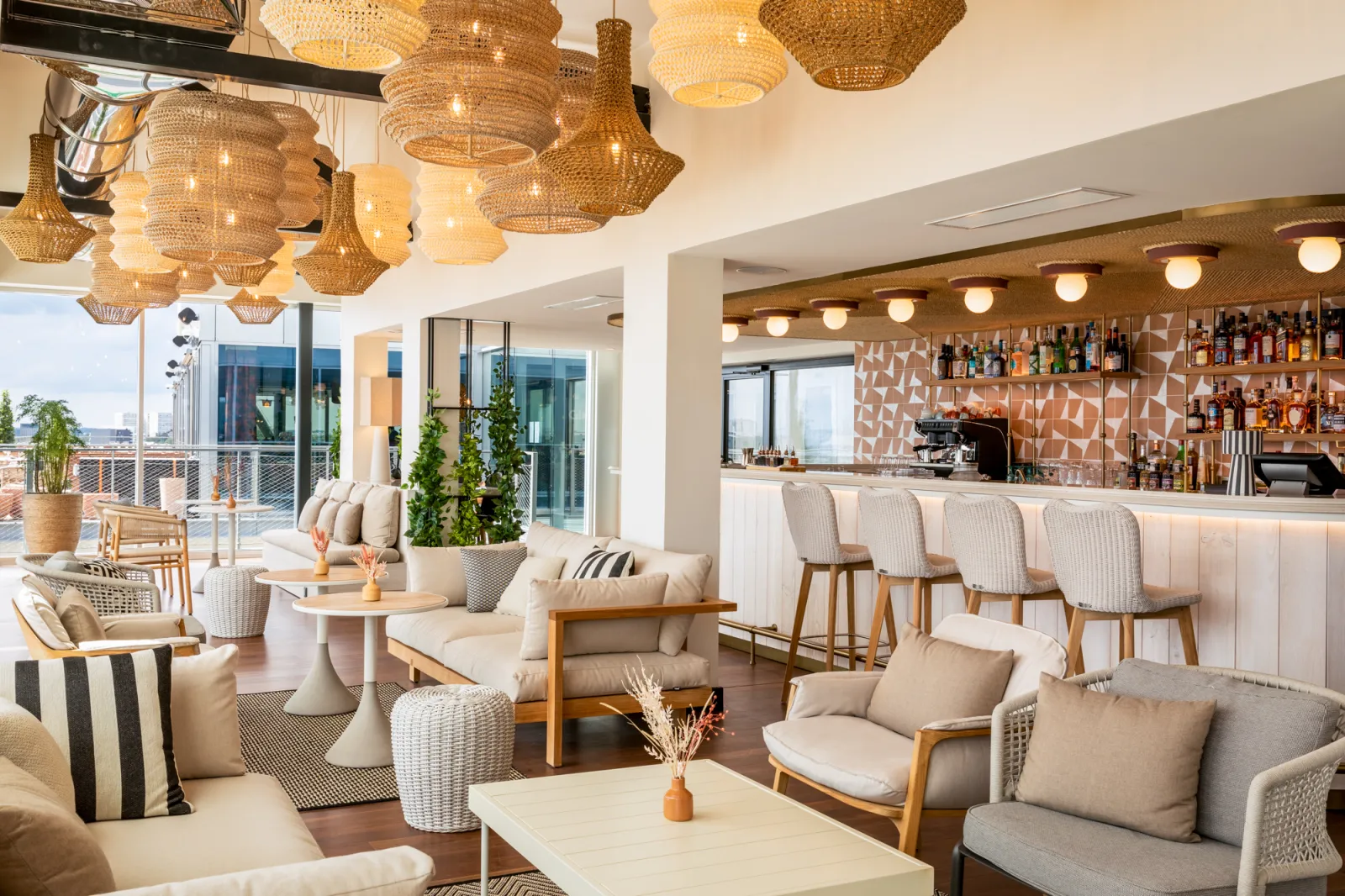 Un élégant bar-lounge avec plafond orné de lustres en rotin, mobilier beige et bois clair, bar bien approvisionné en arrière-plan, et terrasse vitrée surplombant Bordeaux