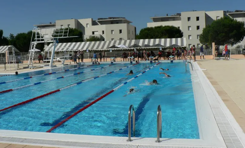 Vue d'une piscine municipale olympique avec plusieurs nageurs dans les couloirs délimités par des cordes rouges et blanches, entourée de spectateurs et d'immeubles résidentiels modernes en arrière-plan à Floirac, près de Bordeaux.