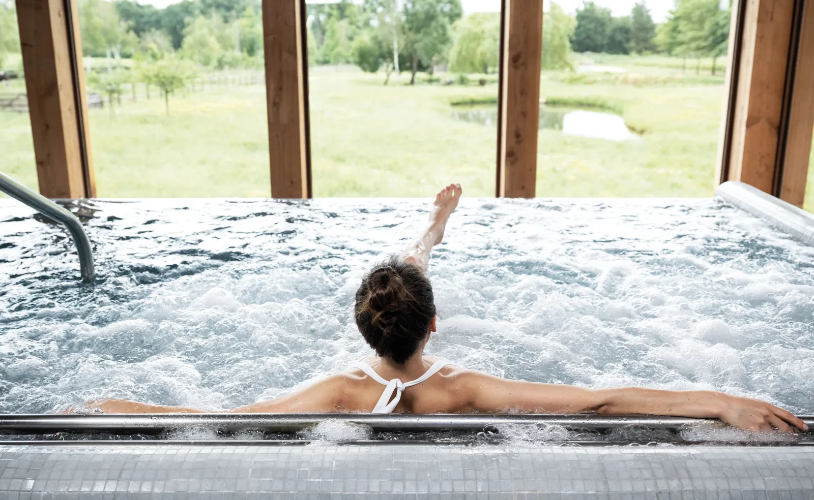 Une femme relaxe dans un bain à remous avec vue panoramique sur les vignobles et la nature environnante à travers les grandes baies vitrées du spa.