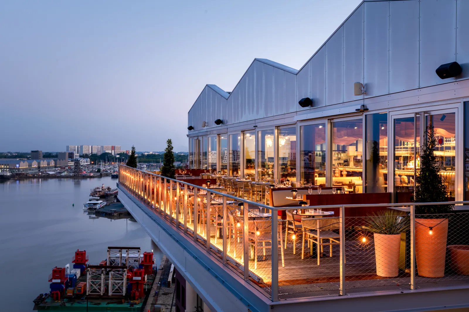 Restaurant moderne en bord de Garonne à Bordeaux au crépuscule, avec terrasse éclairée, baies vitrées panoramiques et vue sur le port fluvial