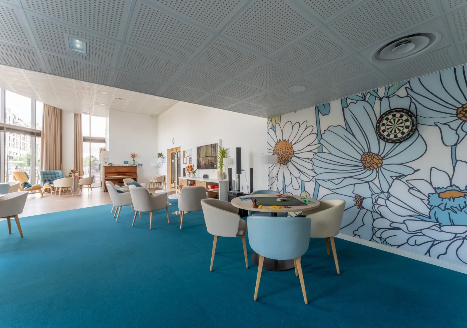 Espace commun lumineux d'une résidence seniors à Bordeaux avec sol bleu, fauteuils modernes, mur orné d'une fresque florale, plafond acoustique et mobilier contemporain.