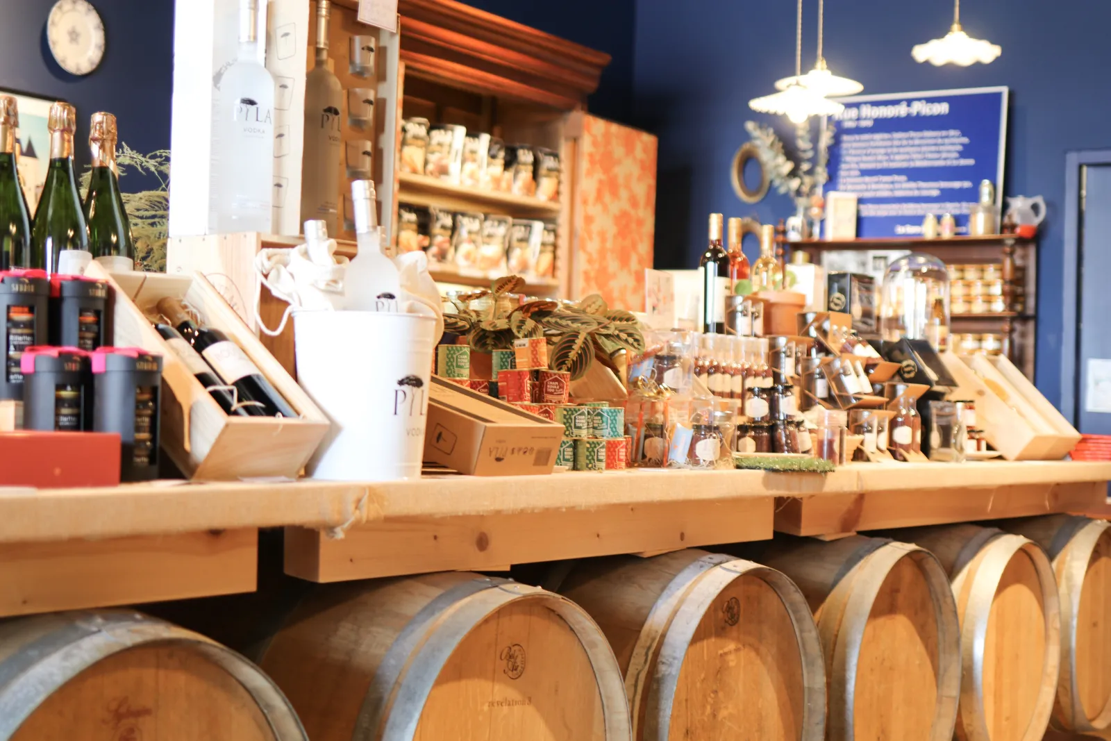 Intérieur d'une échoppe bordelaise avec comptoir en bois, tonneaux de vin en premier plan, étagères remplies de bouteilles et produits locaux, ambiance chaleureuse avec murs bleus et éclairage doré.