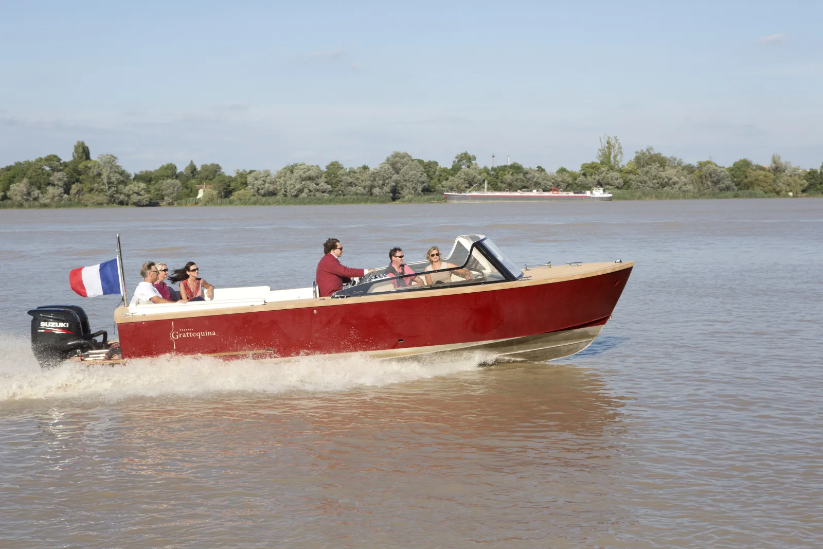 Le bateau taxi Grattequina rouge et blanc navigue sur la Garonne à Bordeaux avec des passagers à bord, un drapeau français flottant sur le côté, et la forêt fluviale en arrière-plan.