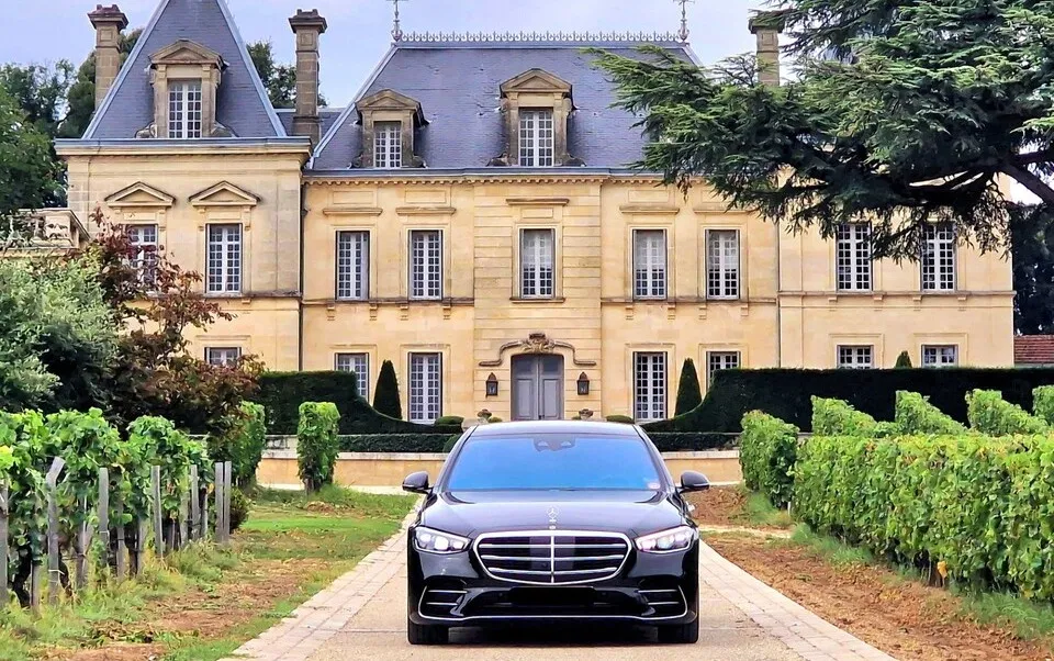 Voiture de luxe noire stationnée sur une allée de vignoble face à un château bordelais en pierre de taille avec toit ardoisé.