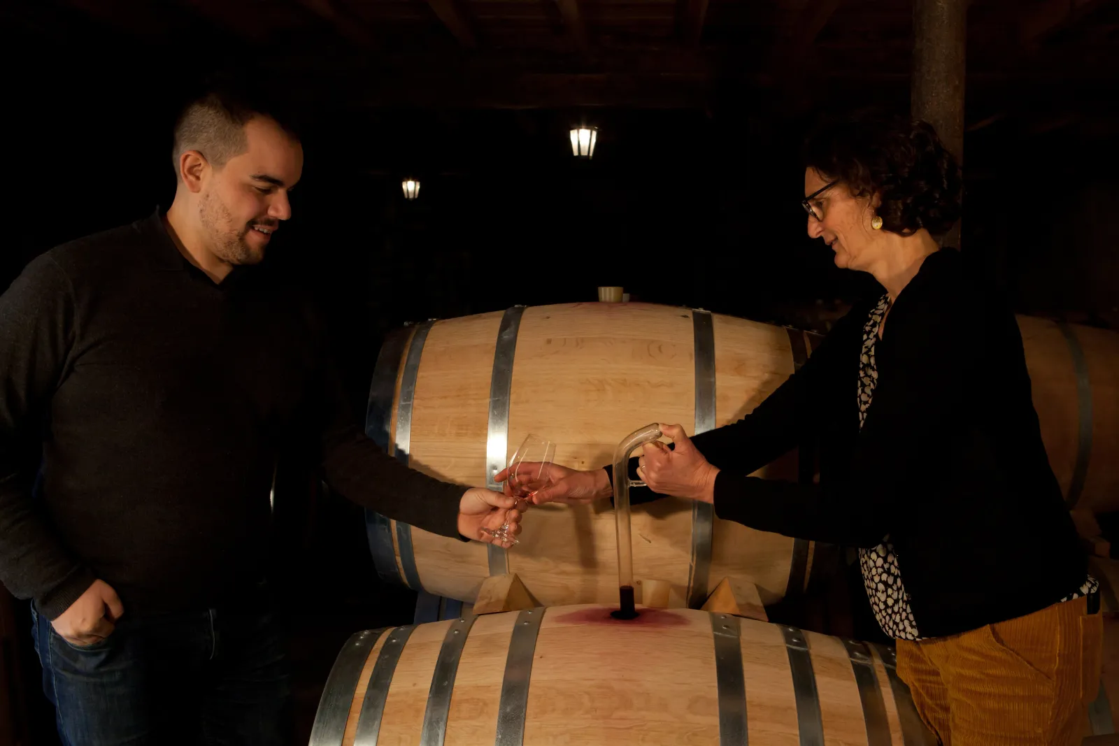 Deux personnes dégustant du vin rouge devant des tonneaux de chêne dans une cave, éclairées par des lanternes murales la nuit.
