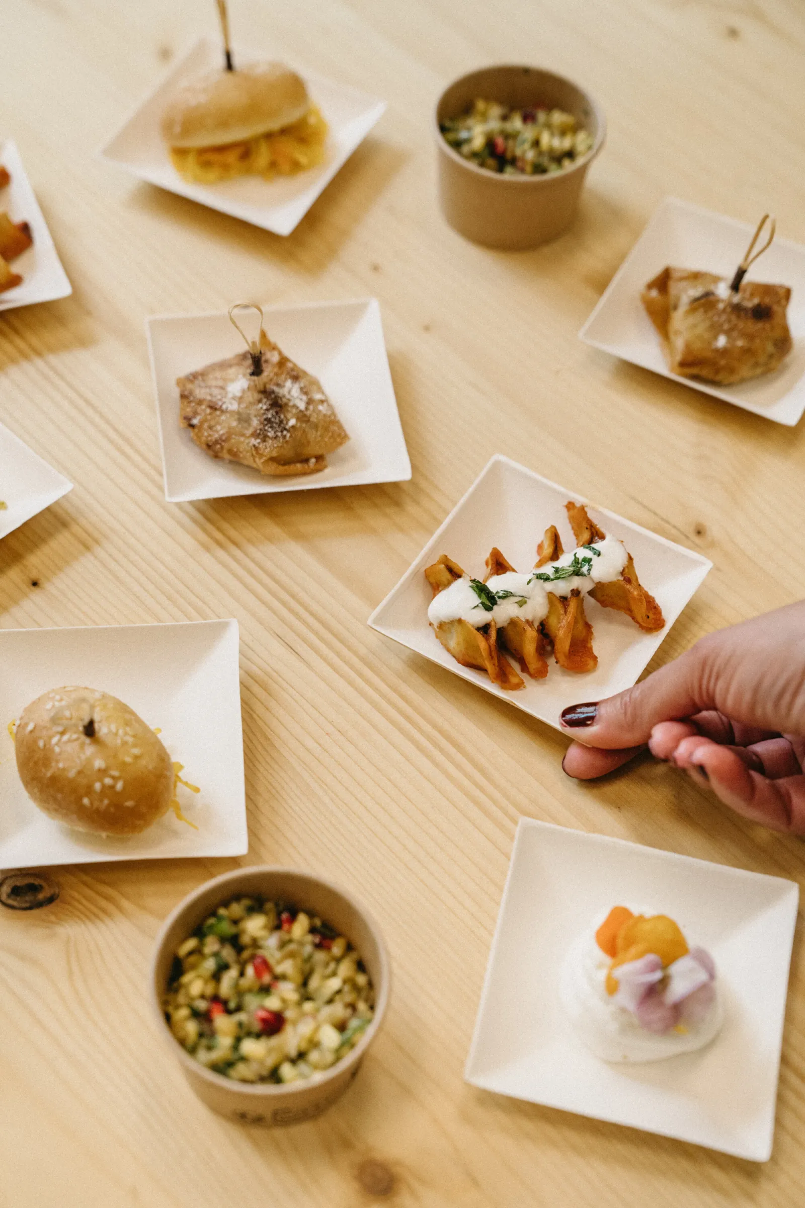 Assortiment de petits plats du restaurant Marie Curry de Bordeaux : samossas, beignets, curry et dessert aux fruits, présentés sur des assiettes blanches et plateaux en bois clair autour d'une main qui saisit un plat.