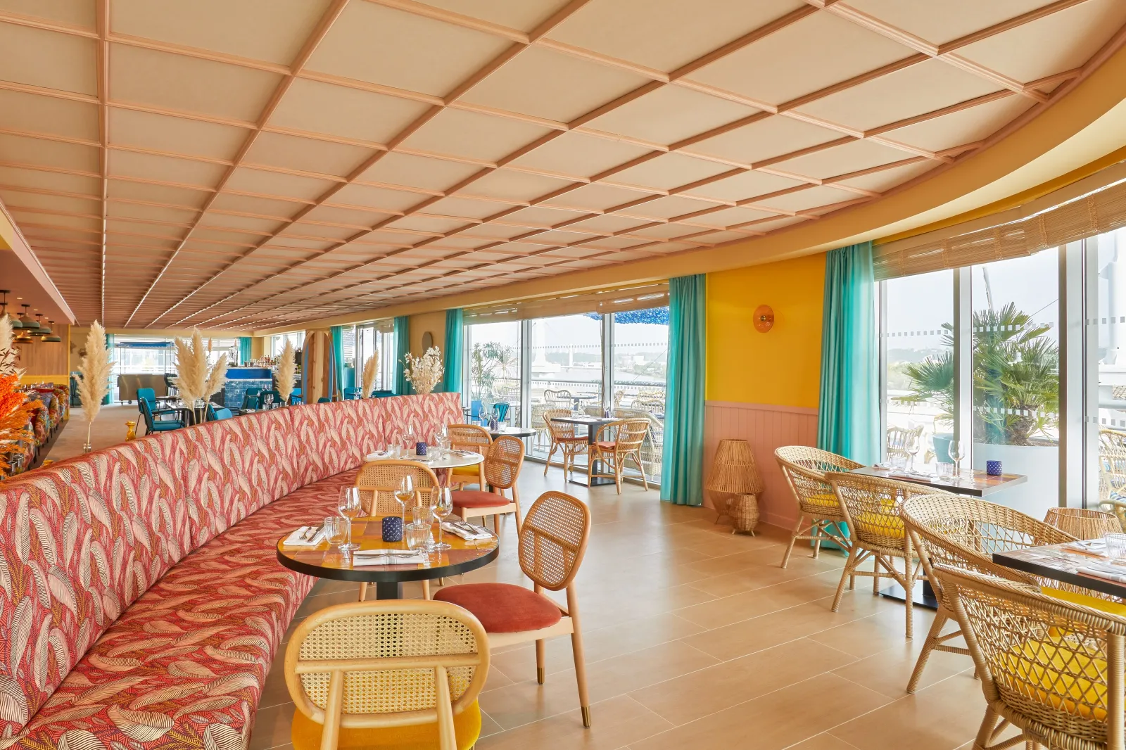 Intérieur moderne d'un restaurant avec plafond en claustras beiges, mobilier en rotin naturel, canapé corail à motifs géométriques, grandes baies vitrées donnant sur un port fluvial, décoration colorée jaune et turquoise, terrasse visible à l'extérieur.