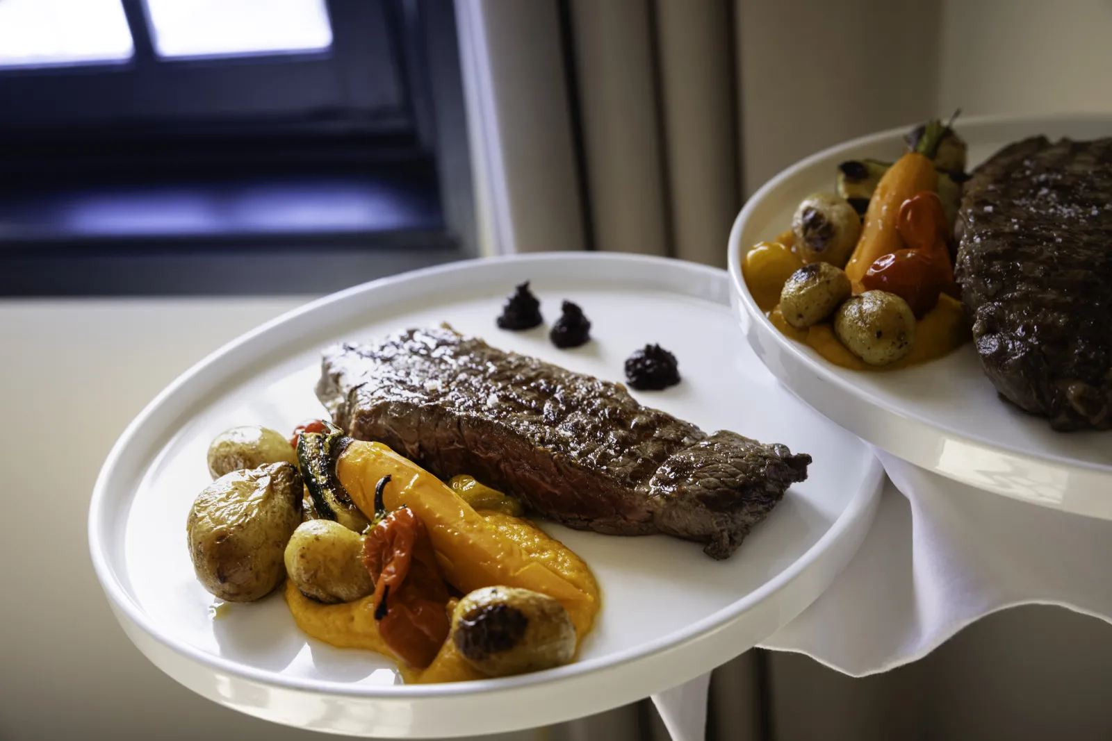 Deux assiettes de viande grillée avec légumes rôtis (poivrons jaunes, champignons et pommes de terre) garnies de baies noires, présentées à L'Atelier de Candale à Bordeaux.