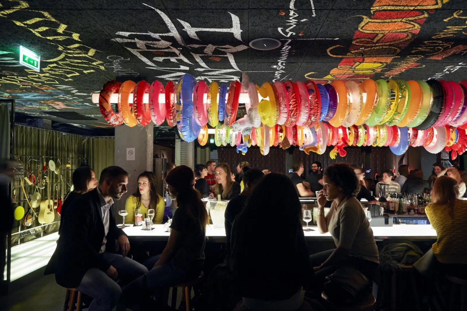 Bar animé du Mama Shelter à Bordeaux avec des clients assis au comptoir illuminé, décoration colorée de bracelets suspendus au plafond et graffitis en arrière-plan.