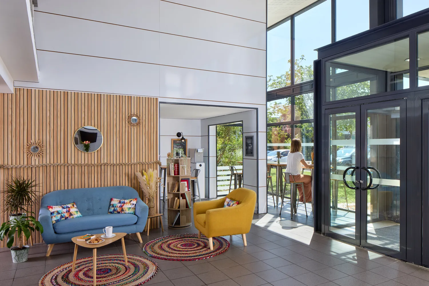 Salon moderne et lumineux avec canapé bleu, fauteuil jaune, tapis colorés et baie vitrée donnant sur une terrasse ensoleillée, murs en bois clair et blanc avec mobilier scandinave.