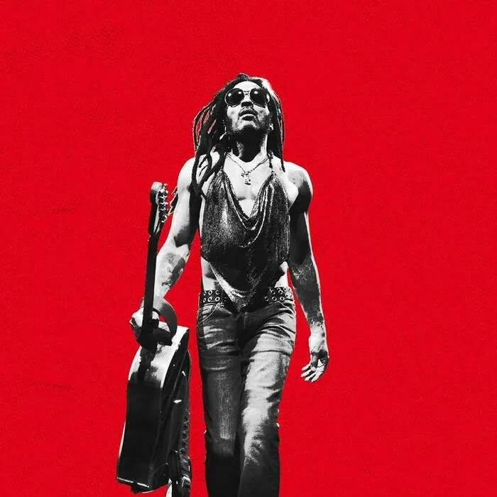 Lenny Kravitz en concert à l'Arkéa Arena de Bordeaux, portant des lunettes de soleil et tenant une guitare, sur fond rouge éclatant.