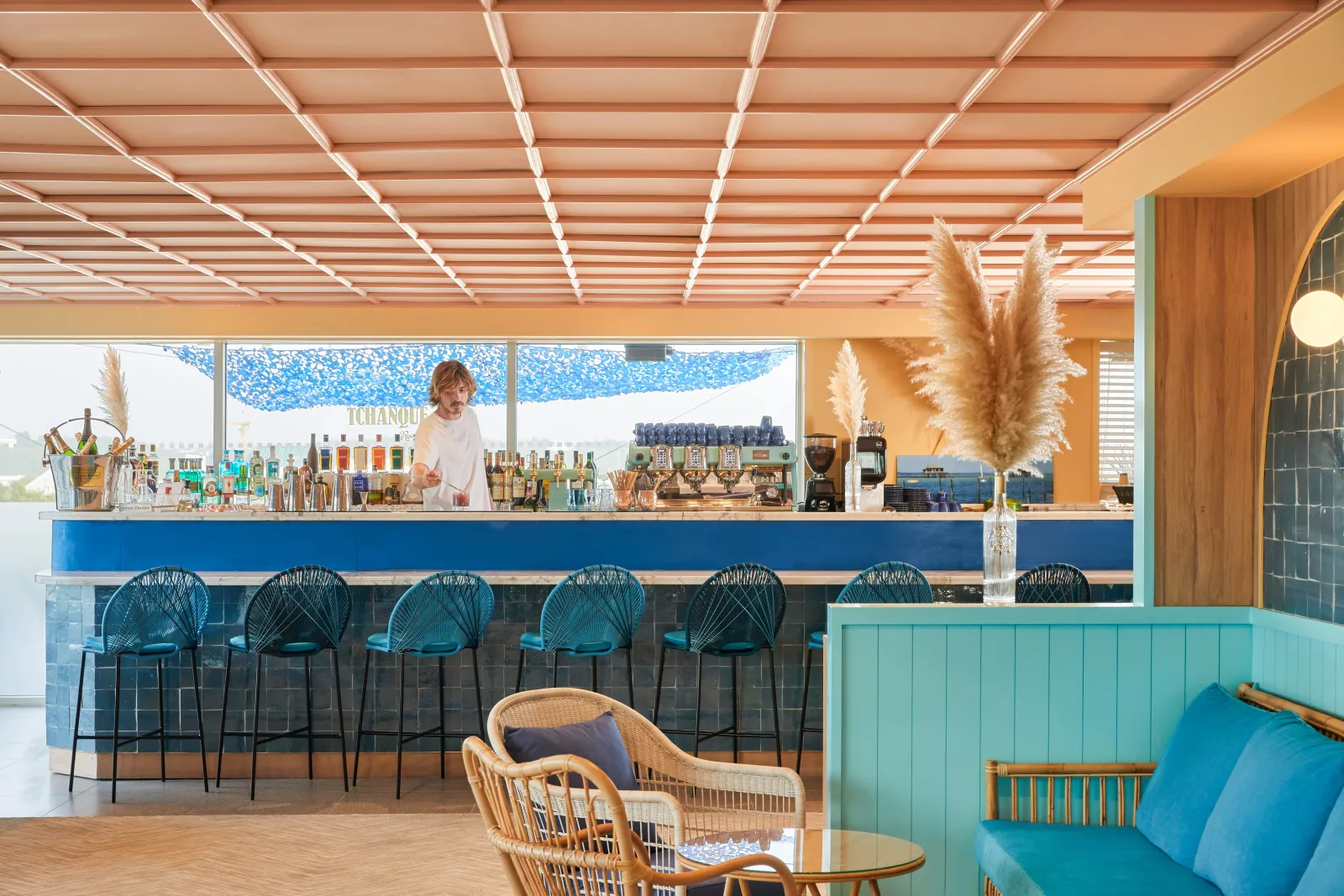 Bar Tchanqué à Bordeaux avec comptoir bleu, tabourets turquoise, plafond à lattes LED, bouteilles de spiritueux, pampas et mobilier en rotin.