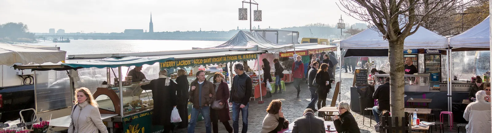 Marché en plein air au quai des Chartrons à Bordeaux avec des étals de vendeurs, des clients flânant entre les stands, et la Garonne en arrière-plan avec la ville visible de l'autre côté du fleuve.